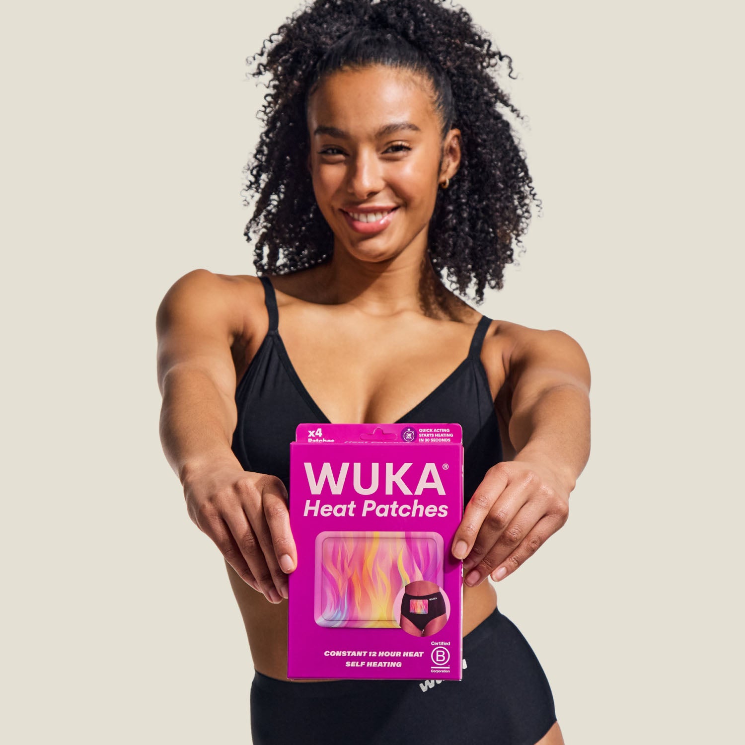 Smiling model in black underwear holding a pack of WUKA Heat Patches, designed for period pain relief.