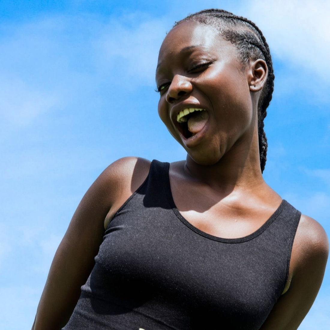 Young woman smiling with playful energy against a bright blue sky, wearing a black tank top and braided hair—radiating confidence and joy outdoors.
